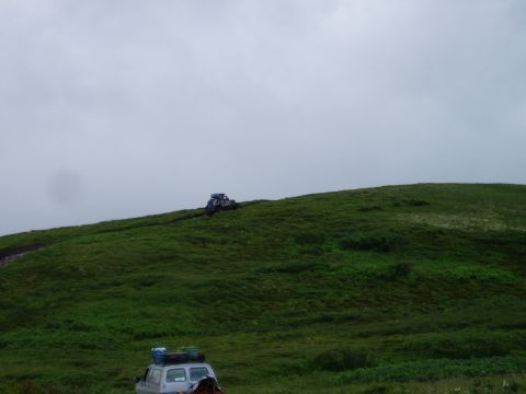 2000 Ford E350 - A white SUV with luggage on its roof is parked on a grassy hill. Another off-road vehicle drives up the hill in the background.