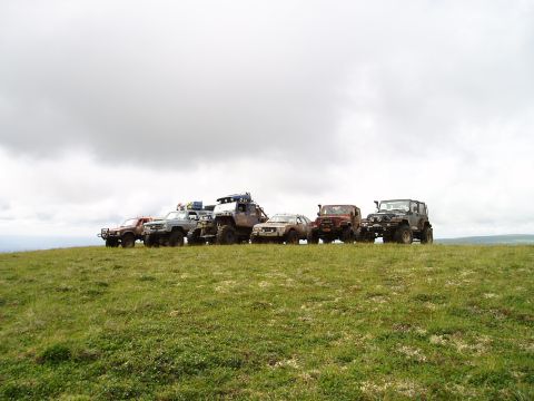 2000 Ford E350 - A line of six off-road modified vehicles are parked on a grassy hill under a cloudy sky. The vehicles are a mix of SUVs and trucks with lar…