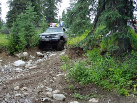 2000 Ford E350 - A lifted, older model Chevrolet pickup truck navigates a rocky, muddy trail through a forest. The truck has off-road tires and accessories…