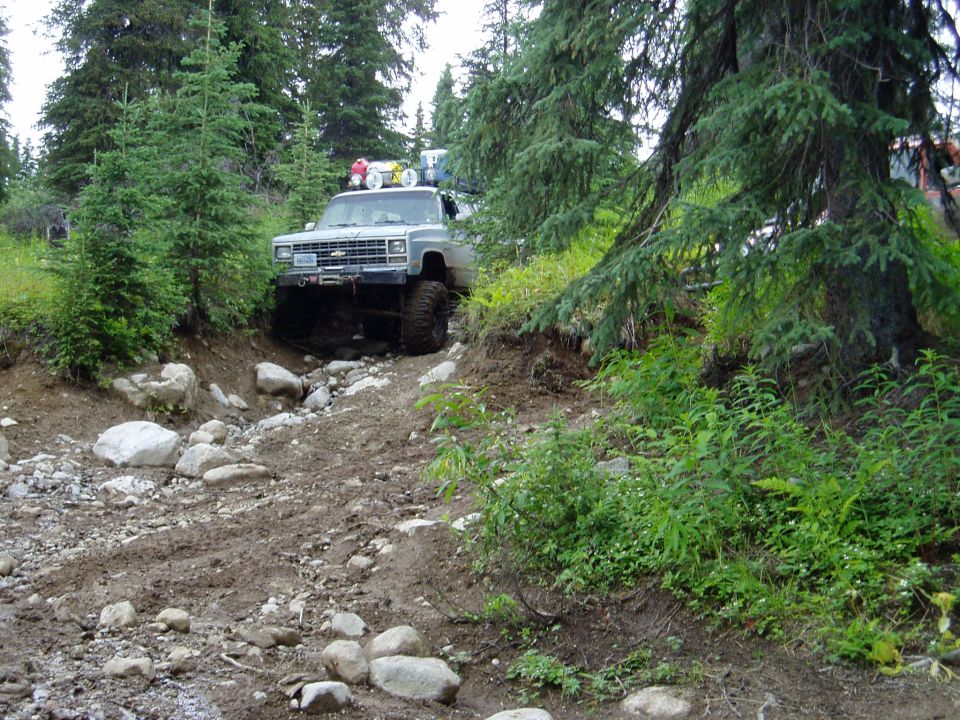 Lifted Chevrolet pickup truck on a muddy, rocky trail in a forest.