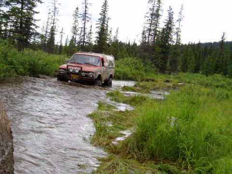 2000 Ford E350 - A red Toyota pickup truck with a white camper shell is driving through a shallow, muddy river in a wooded area.