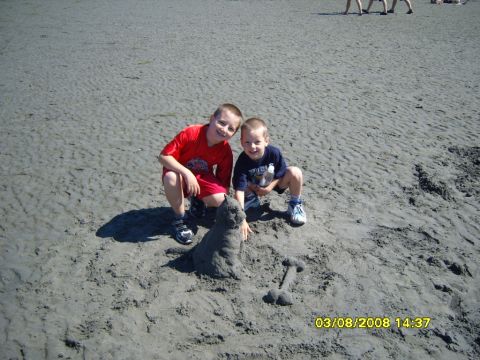1989 Ford Bronco - Two young boys are crouching on a sandy beach next to a sandcastle. The boy on the left is wearing a red t-shirt and red shorts, and the bo…