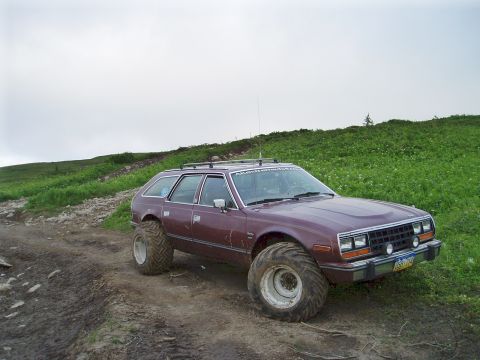 2000 Ford E350 - A maroon AMC Eagle wagon with oversized tires is parked on a muddy dirt road with a grassy hillside in the background.