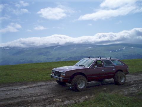 2000 Ford E350 - A lifted, maroon AMC station wagon with large off-road tires sits on a muddy dirt road with a grassy hillside and cloudy mountains in the b…