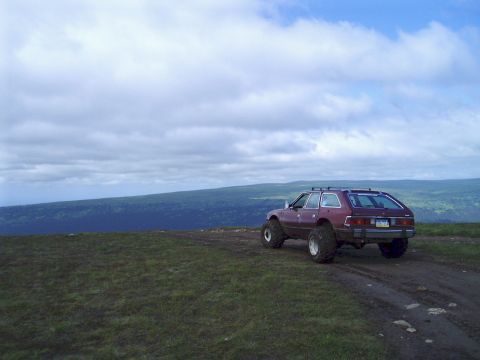 2000 Ford E350 - A lifted, dark red AMC Eagle wagon is parked on a dirt road on a grassy hillside with a vast, tree-covered valley and cloudy sky in the bac…