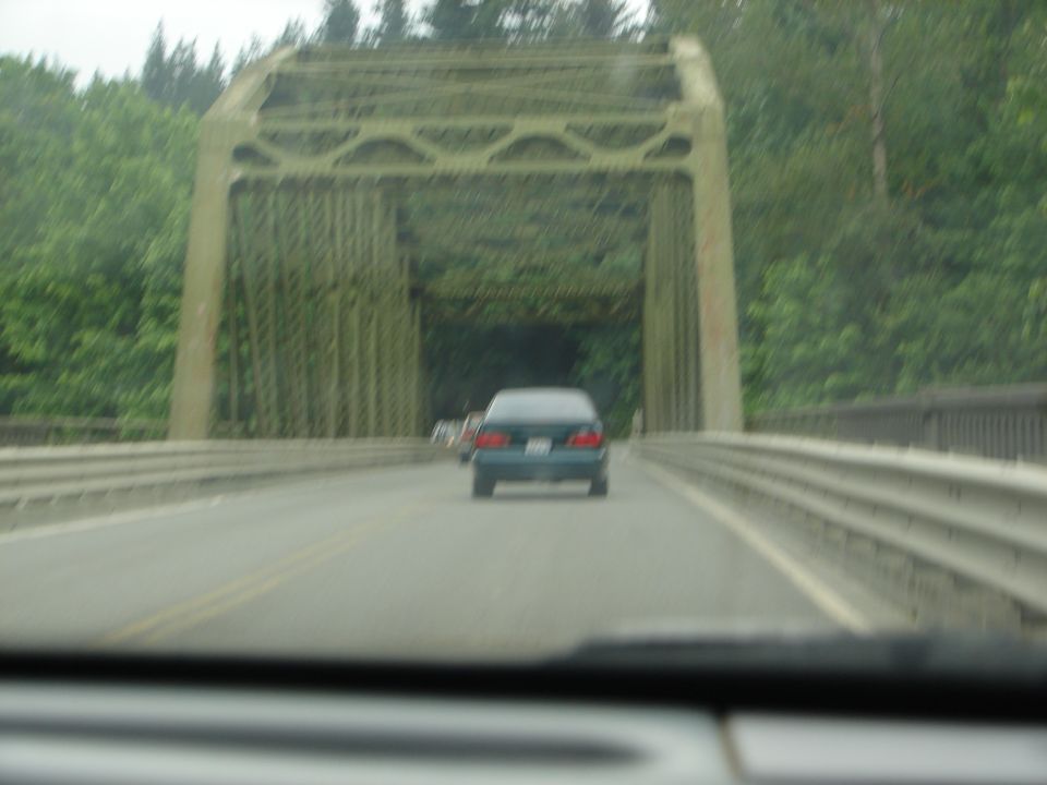 Dark blue sedan driving on a road towards a green truss bridge.