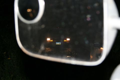 1990 Chevrolet Suburban - A dark sedan is reflected in a side mirror at night, with its taillights illuminated and a license plate visible.