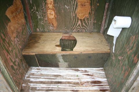 1990 Chevrolet Suburban - The interior of an outhouse shows a wooden toilet seat and a roll of toilet paper mounted on the wall.