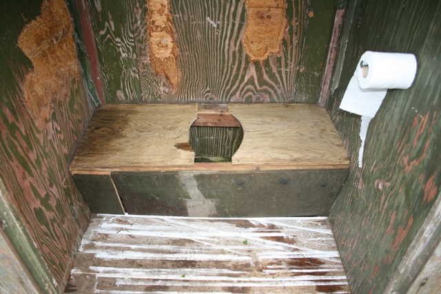 Outhouse interior with wooden toilet seat and roll of toilet paper on the wall