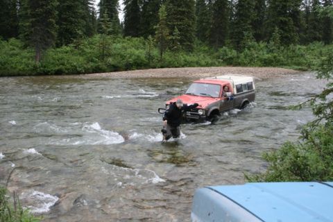 1990 Chevrolet Suburban - A person wades through a river as a red pickup truck with a white camper shell drives through the water.