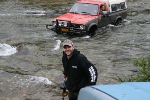 1990 Chevrolet Suburban - A red and brown Toyota pickup truck drives through a shallow river, with two people inside. A man in a black hoodie stands in the foregroun…
