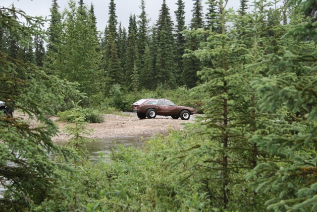 Brown station wagon with white wheels parked by a river in a forest.