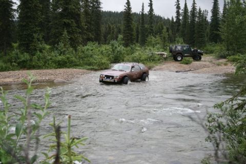 1990 Chevrolet Suburban - A brown station wagon drives through a shallow river with a forest in the background. A green Jeep is parked on the riverbank to the right.
