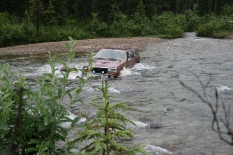 1990 Chevrolet Suburban - A maroon SUV is driving through a shallow, rocky river with trees and brush lining the banks.