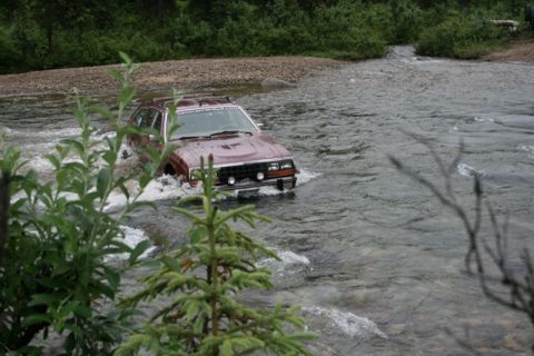 1990 Chevrolet Suburban - A maroon station wagon is driving through a shallow river, with trees and foliage in the background. The vehicle's tires are submerged in t…