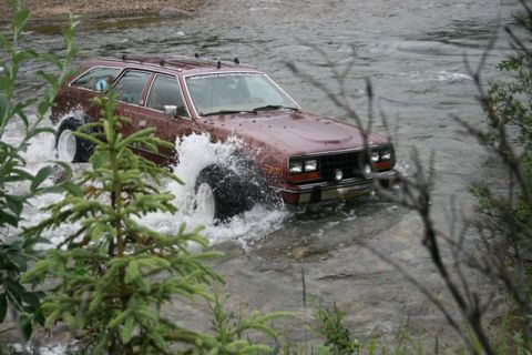 1990 Chevrolet Suburban - A maroon station wagon with large tires drives through a shallow river, creating a large splash. The vehicle has a roof rack and a sticker…