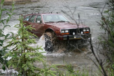 1990 Chevrolet Suburban - A maroon station wagon is driving through a shallow river, kicking up water. The vehicle appears to be modified for off-road use with large…