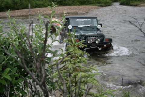 1990 Chevrolet Suburban - A dark green Jeep Wrangler is driving through a shallow river, kicking up water. The vehicle has multiple aftermarket lights mounted on the…