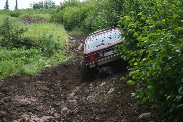 Maroon station wagon driving down a steep, muddy off-road trail.
