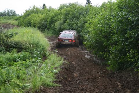 1990 Chevrolet Suburban - A maroon station wagon is driving on a muddy, rutted dirt road surrounded by lush green foliage. The vehicle is seen from the rear as it na…