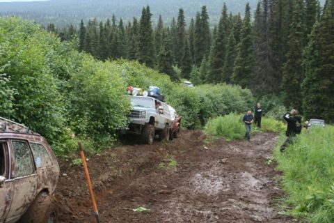 1990 Chevrolet Suburban - A lifted, modified Chevrolet Suburban navigates a very muddy, rutted off-road trail surrounded by dense green foliage and tall pine trees.…