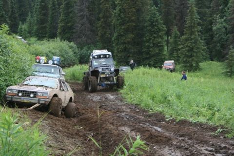 1990 Chevrolet Suburban - Several off-road vehicles are navigating a muddy trail through a forest with tall green trees. A light-colored AMC Pacer is stuck in the mu…