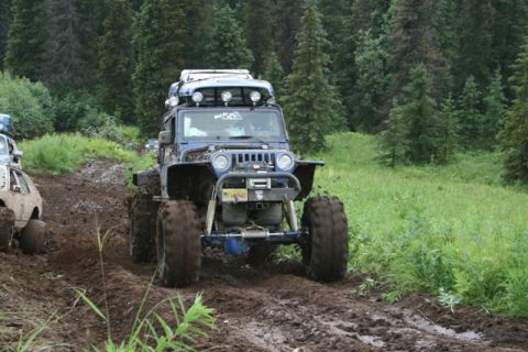1990 Chevrolet Suburban - A modified blue Jeep Wrangler is driving through a muddy trail in a forest. Another off-road vehicle is visible to the left.