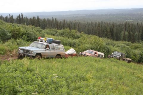 1990 Chevrolet Suburban - A light blue Chevrolet Suburban, loaded with gear on its roof rack, navigates a muddy, overgrown trail, followed by two other off-road vehi…