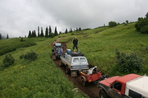1990 Chevrolet Suburban - A convoy of off-road vehicles, including a light blue Chevrolet Suburban towing a trailer, navigates a muddy trail through a grassy, treed…