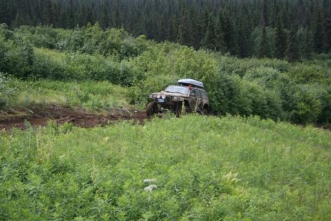 1990 Chevrolet Suburban - A lifted, mud-covered Jeep Cherokee with a roof-top carrier navigates a muddy, overgrown trail surrounded by dense forest.