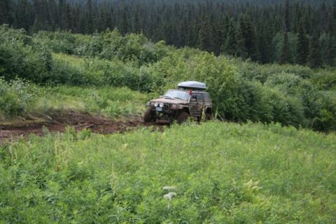 1990 Chevrolet Suburban - A muddy, dark-colored SUV with a roof-top cargo carrier navigates a dirt path through a grassy, wooded area. The vehicle is angled towards…