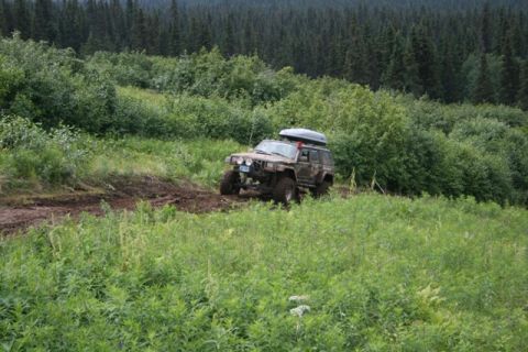 1990 Chevrolet Suburban - A muddy, lifted Jeep Cherokee XJ with a roof-top cargo box navigates a rutted dirt trail through a grassy, wooded hillside.