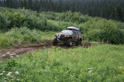 1990 Chevrolet Suburban - A lifted, muddy Jeep Cherokee XJ with a roof-top cargo box drives off-road on a dirt path surrounded by lush green vegetation and a dense f…