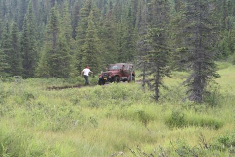 1990 Chevrolet Suburban - A person in a red hat and white shirt walks near a reddish-brown Jeep Wrangler that is partially stuck in mud in a wooded area.