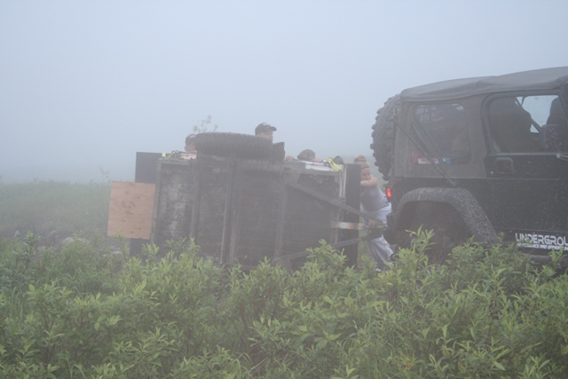 People trying to right a tipped-over trailer behind a black Jeep Wrangler in foggy conditions.