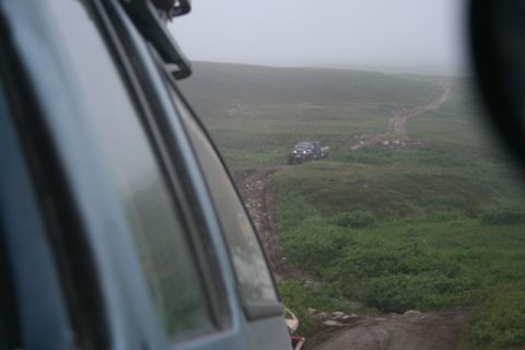 1990 Chevrolet Suburban - View from inside a vehicle looking out at a dirt track leading up a grassy hill. Two other dark-colored SUVs are visible on the track in th…