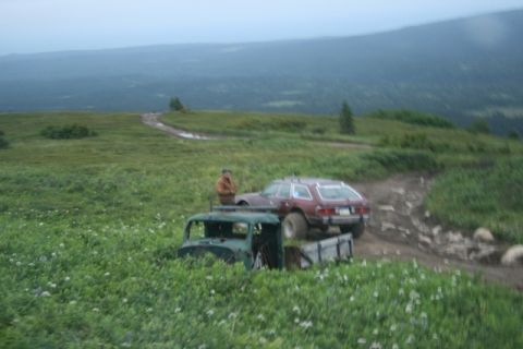1990 Chevrolet Suburban - A man in a brown jacket stands next to a vintage green truck cab and bed in a grassy field, with a maroon station wagon parked on a dirt ro…