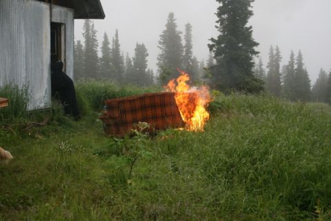 1990 Chevrolet Suburban - A couch is engulfed in flames in a field near a rustic building, with trees in the background.