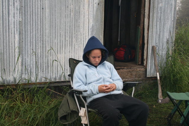 Person in blue hoodie and black pants sits in a camping chair outside a metal building