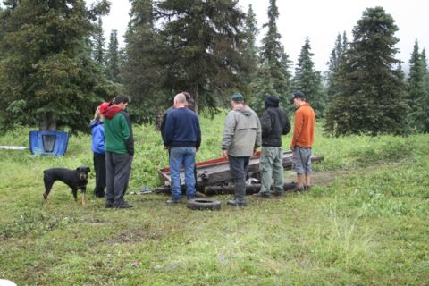 1990 Chevrolet Suburban - A group of people and a dog examine a damaged vehicle chassis in a grassy, wooded area.
