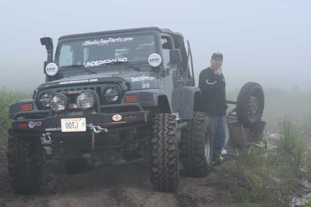 Green Jeep Wrangler with snorkel, big tires, and aftermarket bumper. Guy in jeans and black shirt stands next to it