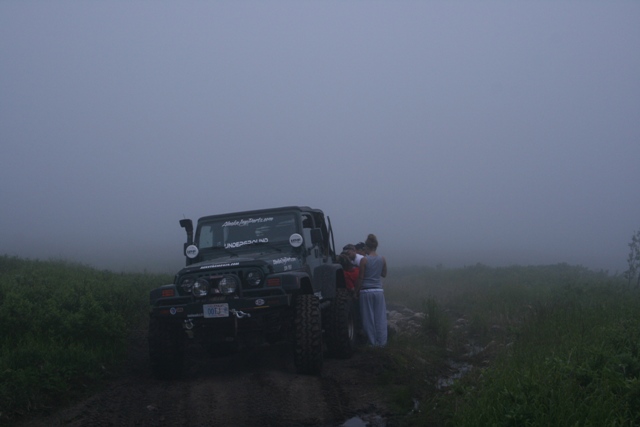 Jeep Wrangler on a muddy trail in fog, with several people standing nearby