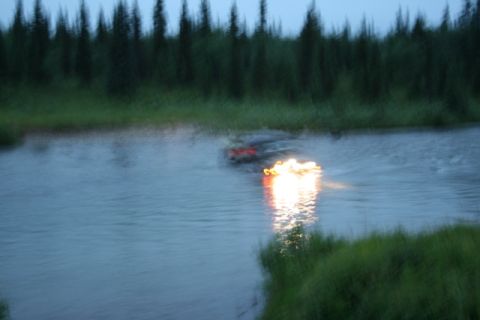 1990 Chevrolet Suburban - A dark-colored vehicle, possibly an SUV, is partially submerged and moving through a body of water at dusk. Bright lights are visible on th…