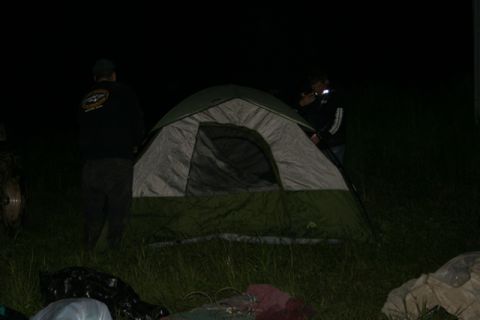 1990 Chevrolet Suburban - Two people are setting up a tent in a grassy area at night. One person stands to the left of the tent, facing away from the camera, while t…