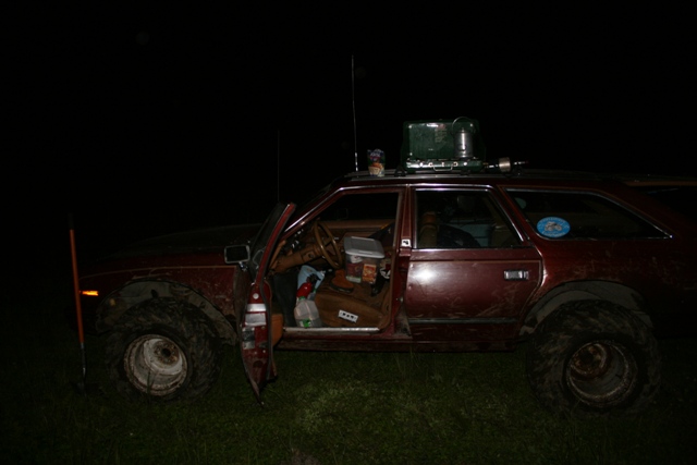 Maroon station wagon with large tires and a stove on the roof, door open at night.