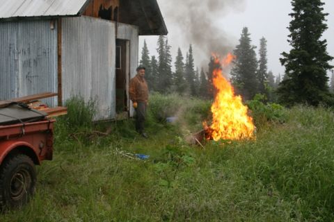 1990 Chevrolet Suburban - A man stands near a burning barrel next to a corrugated metal building. A red trailer with lumber is parked to the left of the building.