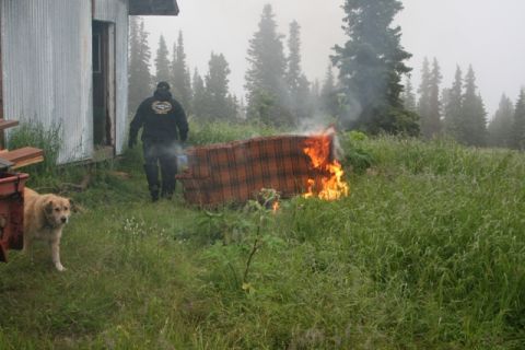 1990 Chevrolet Suburban - A person in a black hoodie stands near a burning couch in a grassy, wooded area with a dog in the foreground.