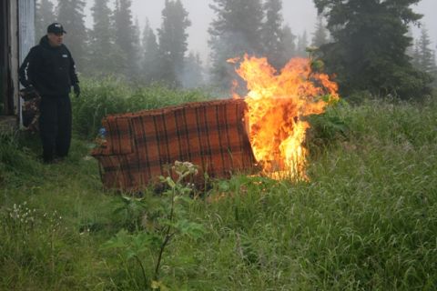 1990 Chevrolet Suburban - A person stands next to a couch that is engulfed in flames in a grassy, wooded area.