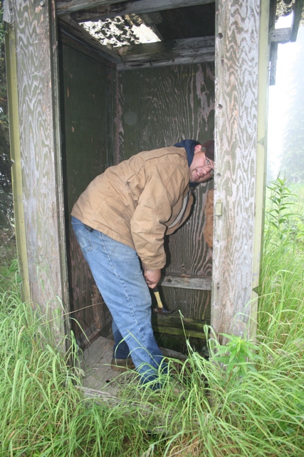 1990 Chevrolet Suburban - A person is inside a dilapidated wooden outhouse, bent over and holding a hammer as if to break something. Tall grass surrounds the structu…