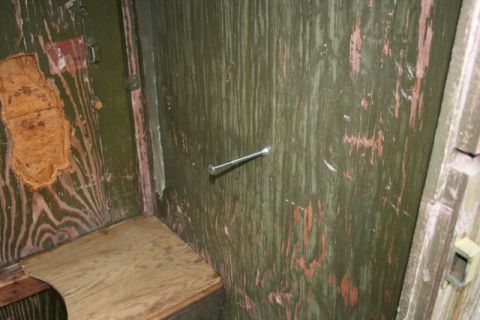 1990 Chevrolet Suburban - The interior of what appears to be a wooden outhouse or shed, showing a wooden seat and a bolt protruding from the green painted wall.